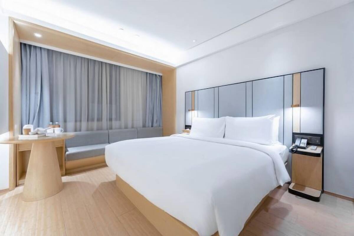 A modern hotel room with a large bed, a bedside table, a desk, and a large window with curtains.