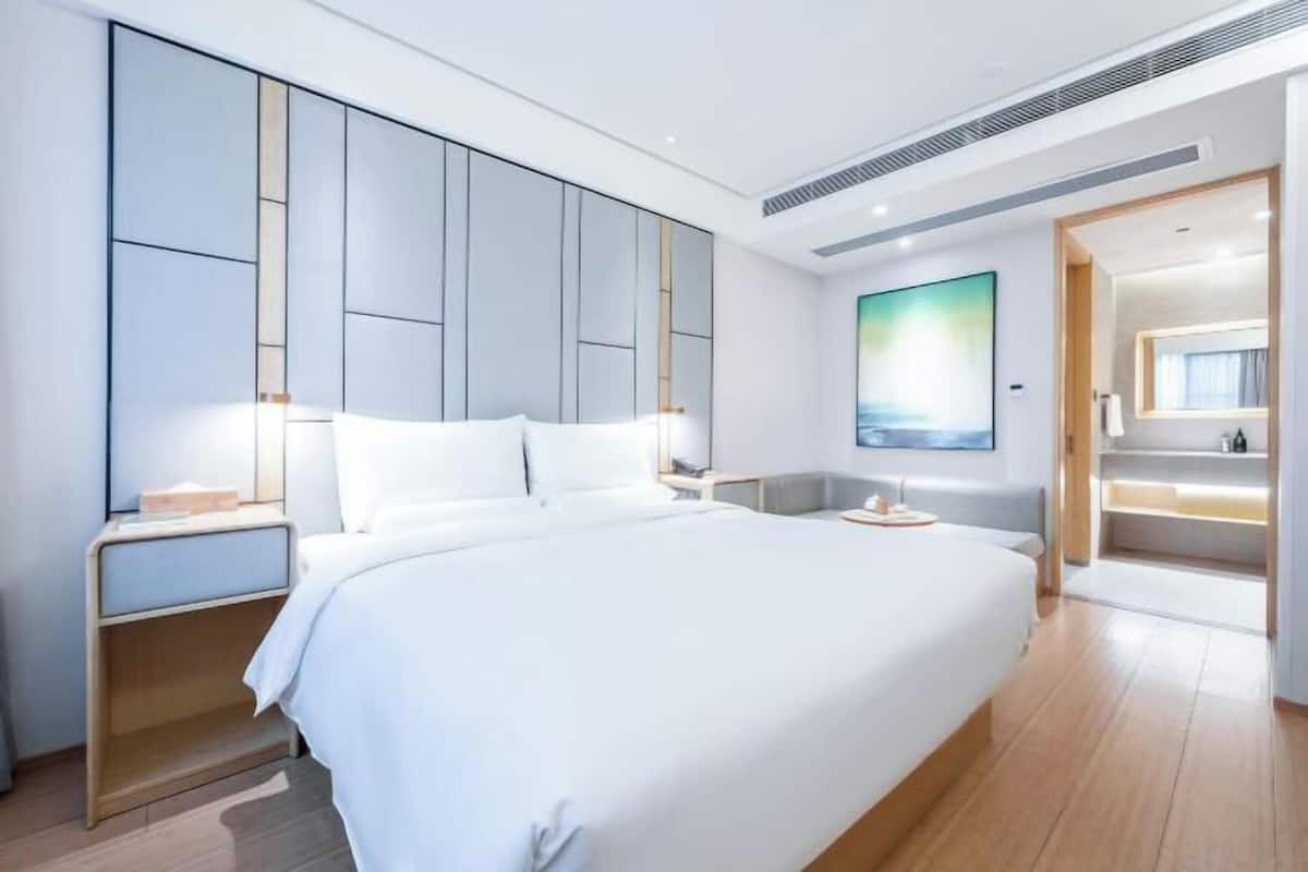 A modern hotel room with a large bed, built-in wardrobe, bedside tables, a painting on the wall, and a bathroom visible through an open door.