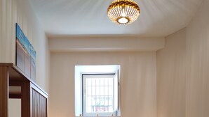 Ein ordentlich bezogenes Bett mit einem gemusterten Überwurf, zwei Nachttischlampen, einem Fenster mit Gitter und einer Pendelleuchte.
