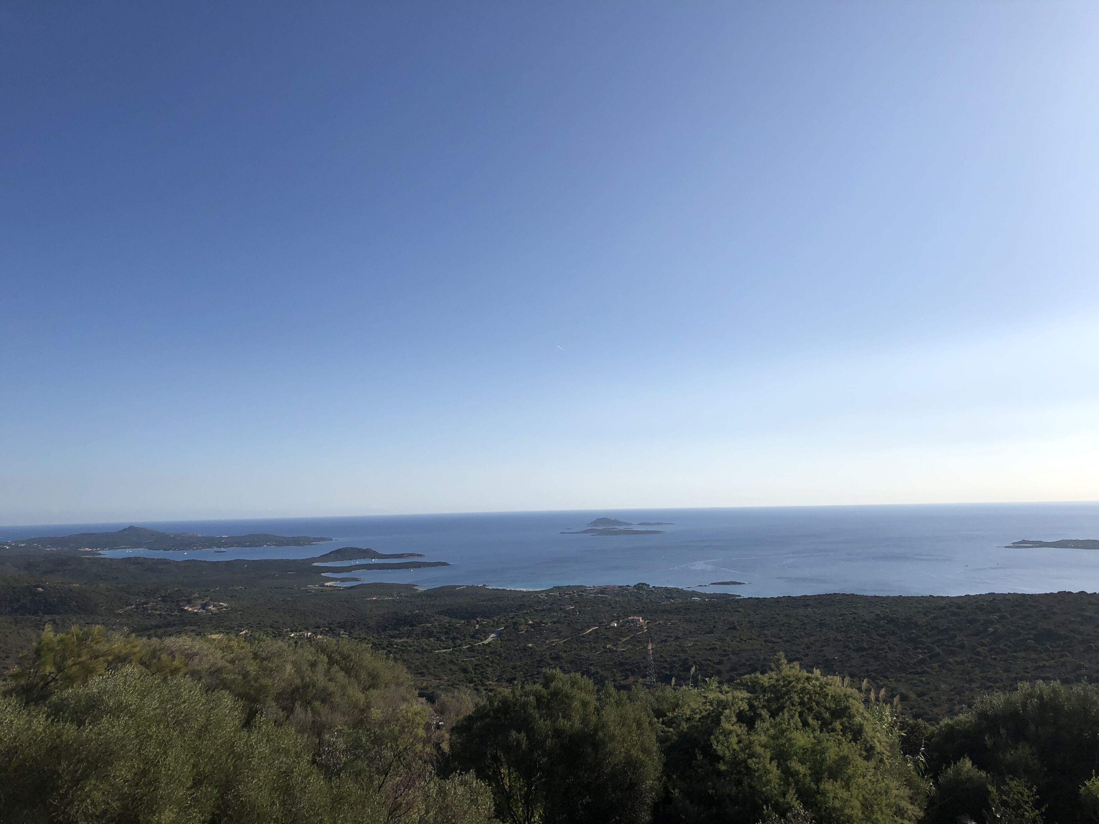 Ett kustlandskap med klar himmel, tät vegetation och en avlägsen utsikt över havet och öarna.