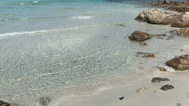 Een strand met helder turquoise water, een rotsachtige kustlijn en een landmassa in de verte.