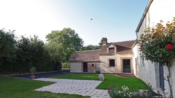 A stone pathway leading to a house with a brick chimney, surrounded by a well-maintained lawn and a small garden with flowers.