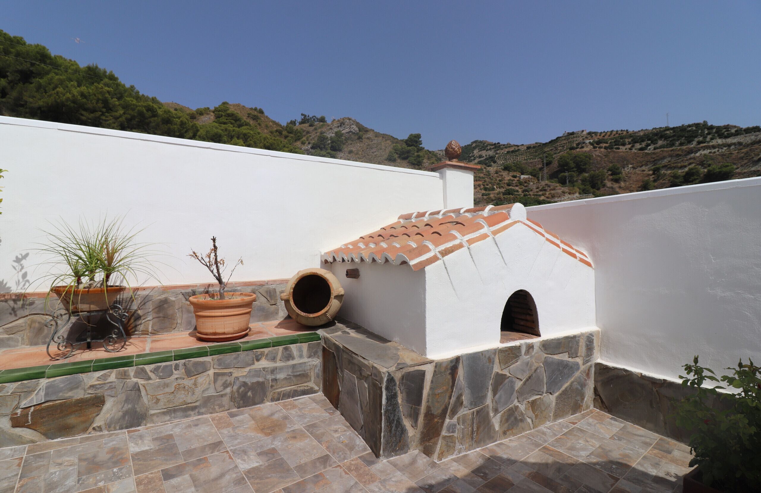 A rooftop terrace with stone flooring, a small structure with a tiled roof, and potted plants.