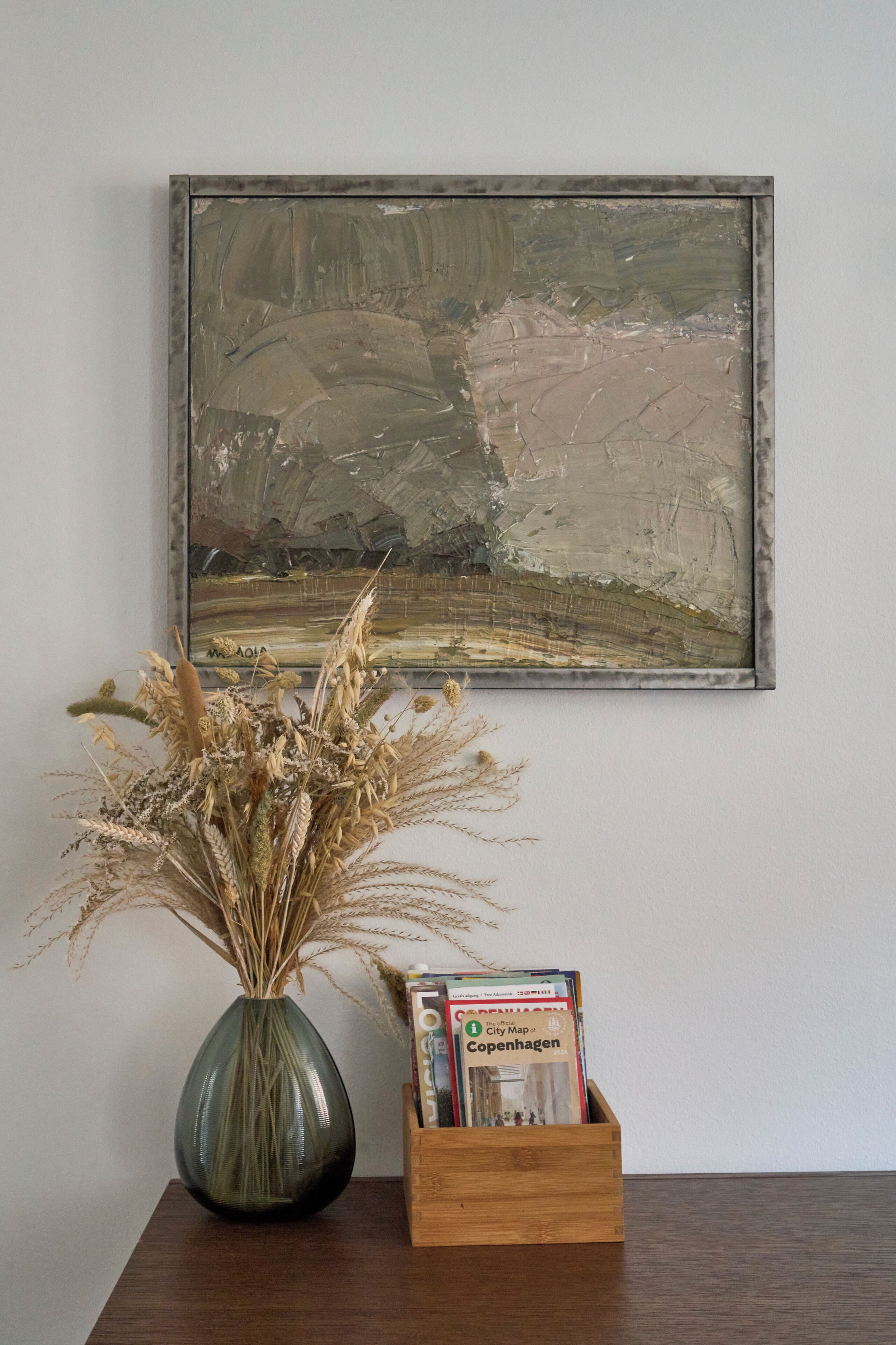 A framed painting, a vase with dried plants, and a wooden box with magazines on a table.