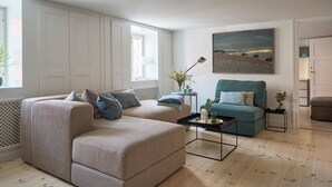 A modern living room with a beige sofa, a blue armchair, and a coffee table. There is a painting on the wall and a window providing natural light. - Perfect location, cosy and stylish and with one of the largest bathrooms i town (København)
