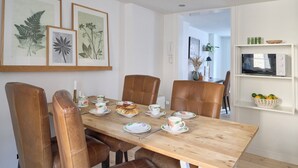 A dining area with a wooden table set for four, surrounded by leather chairs. There are framed botanical prints on the wall, a white shelf with a microwave and a basket of lemons, and a door leading to another room. - Perfect location, cosy and stylish and with one of the largest bathrooms i town (København)