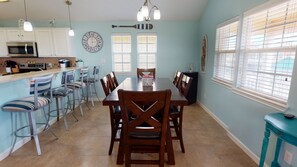Un área de comedor con una mesa y sillas de madera, una cocina con armarios blancos y un reloj, y una ventana con persianas.