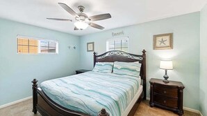 Un dormitorio con cama, mesita de noche, ventilador de techo y ventana con persianas.