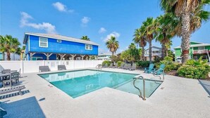 Un área de piscina con un edificio azul, sillones reclinables y palmeras.