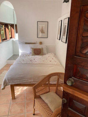 Quarto com uma cama, uma cadeira de vime, uma porta de madeira e uma janela com persianas de madeira.