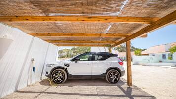 En hvid SUV parkeret under et træbeklædt pergola i et overdækket parkeringsområde.