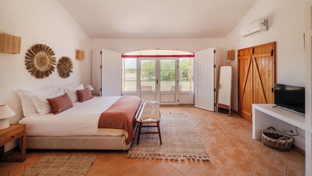 A bedroom with a large bed, a bench, a TV, and a view of the outdoors through a door.