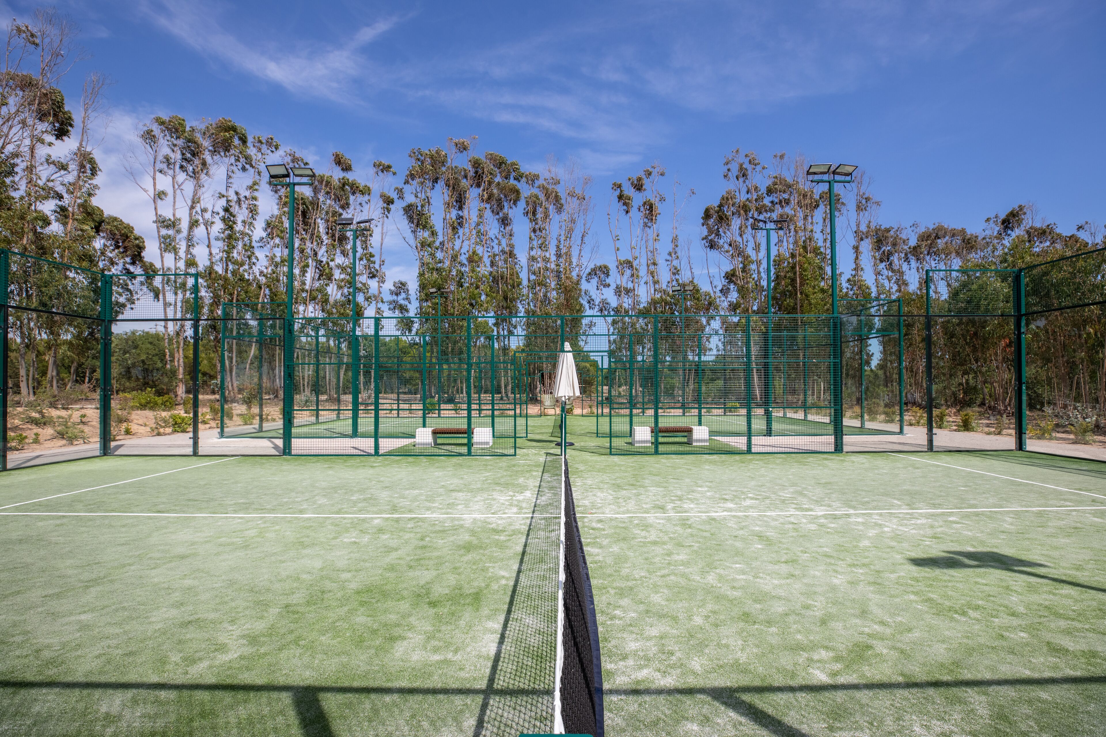 An outdoor tennis court with green fencing, benches, and trees in the background.