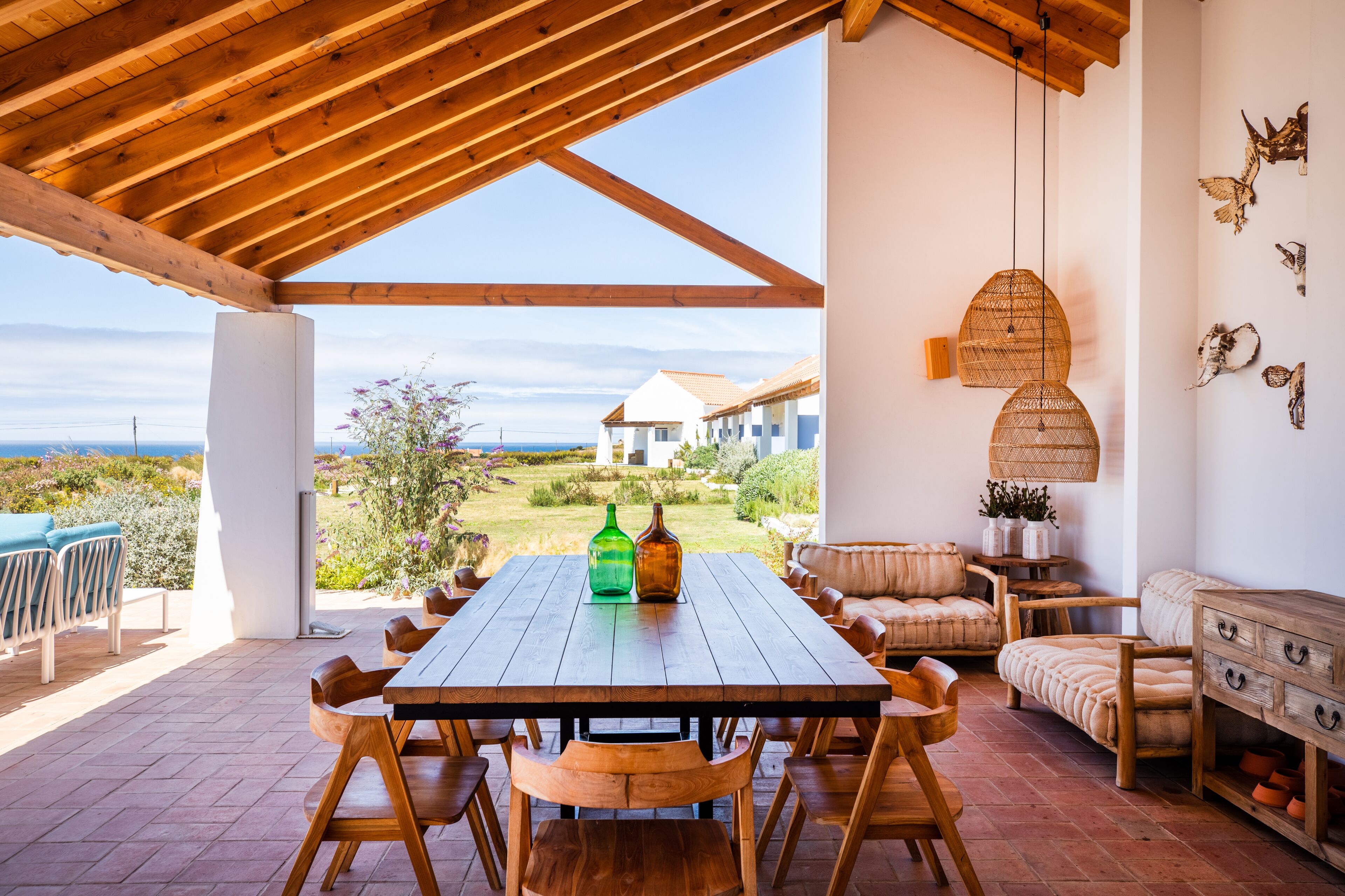 A wooden dining table with chairs, a sofa, and a cabinet in a covered outdoor area with a view of a house and landscape.