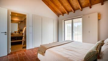 A bedroom with a large bed, a wooden ceiling, a bathroom with a sink and towel rack, and a sliding glass door to the outside.