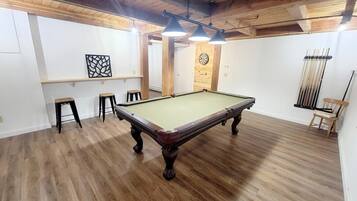 A pool table in a room with wooden floors and a high ceiling. There are stools and a dartboard on the wall.