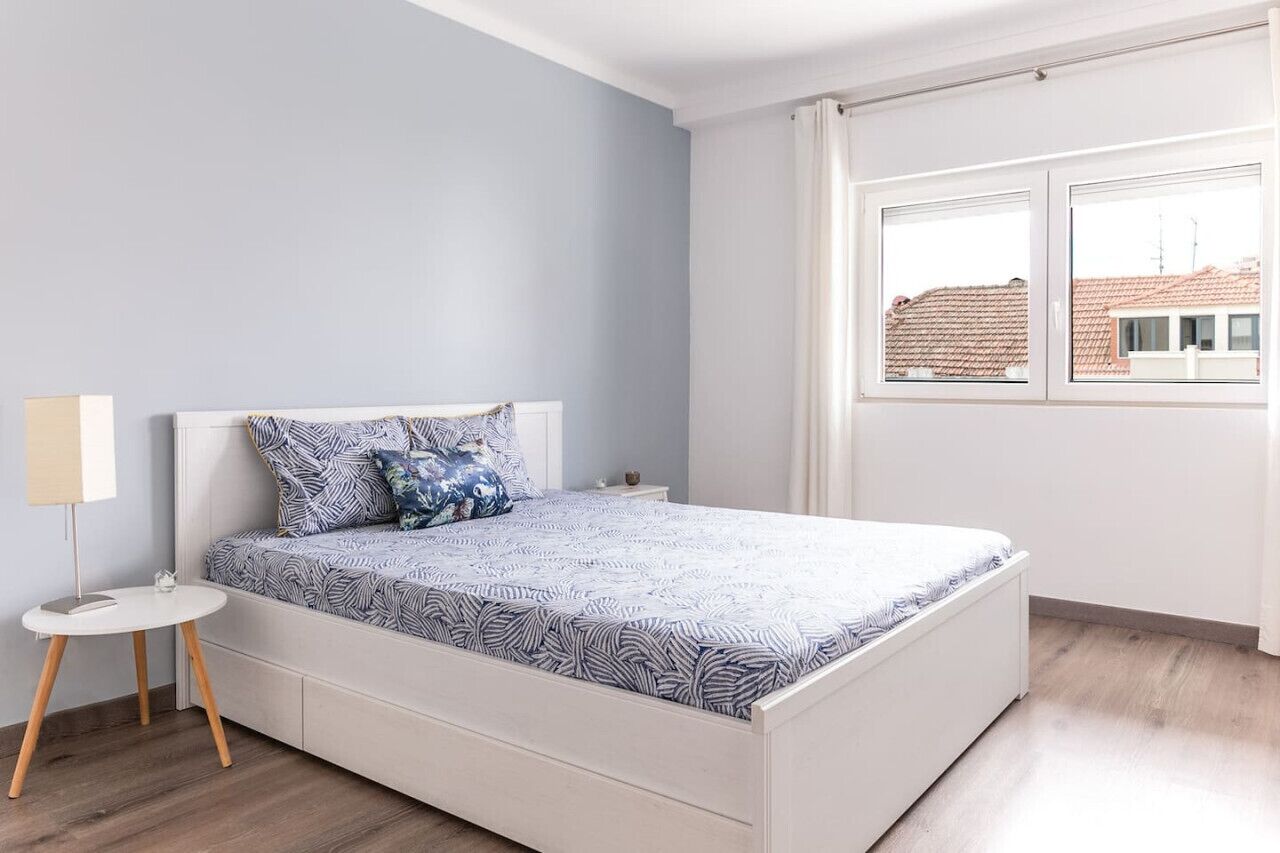 Quarto com cama, mesa de cabeceira pequena, luminária e janela com vista para edifícios.