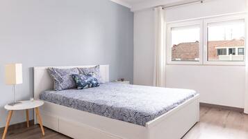 Quarto com cama, mesa de cabeceira pequena, luminária e janela com vista para edifícios.