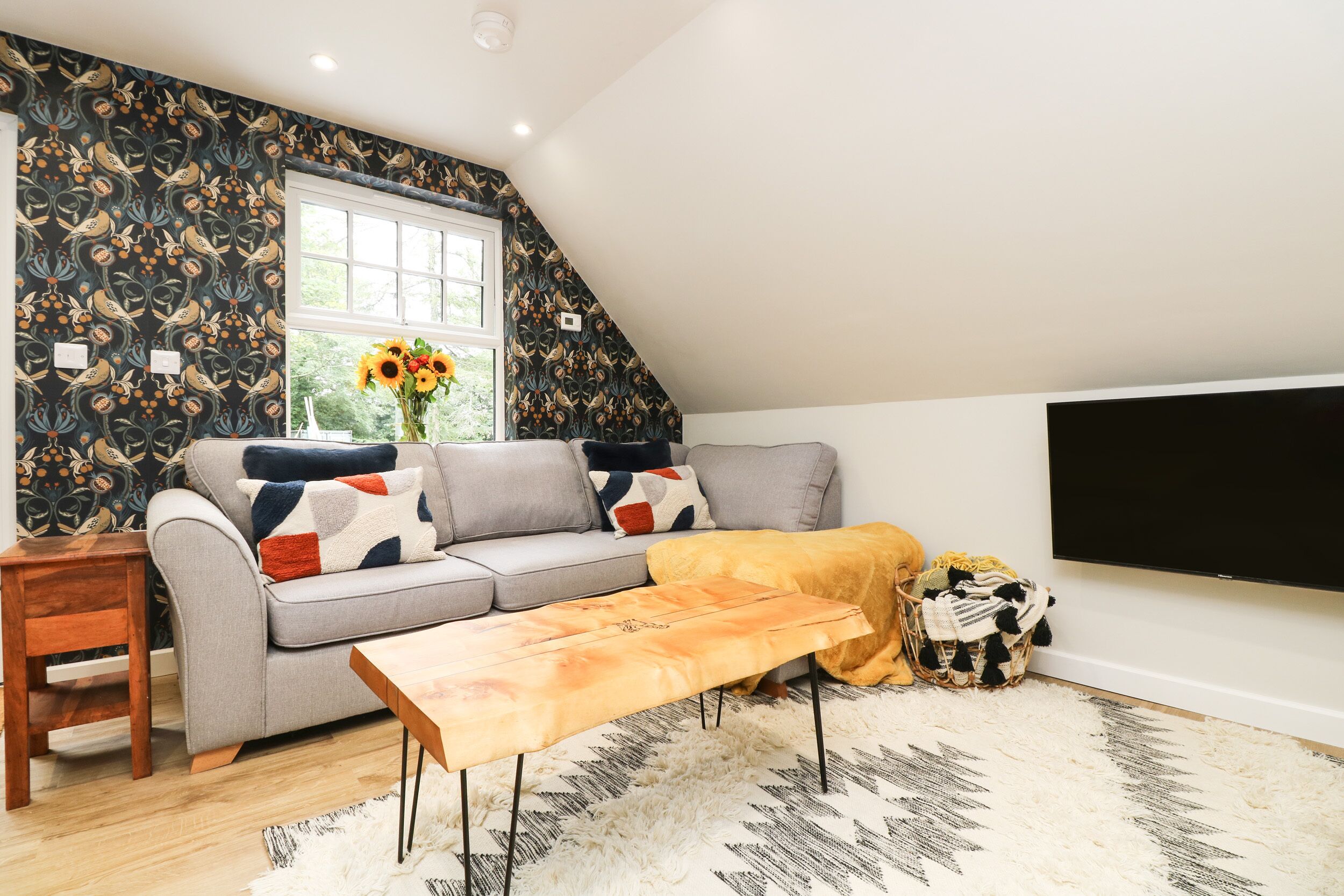 A cozy living room with a grey sofa, a wooden coffee table, and a large window with a view of greenery.