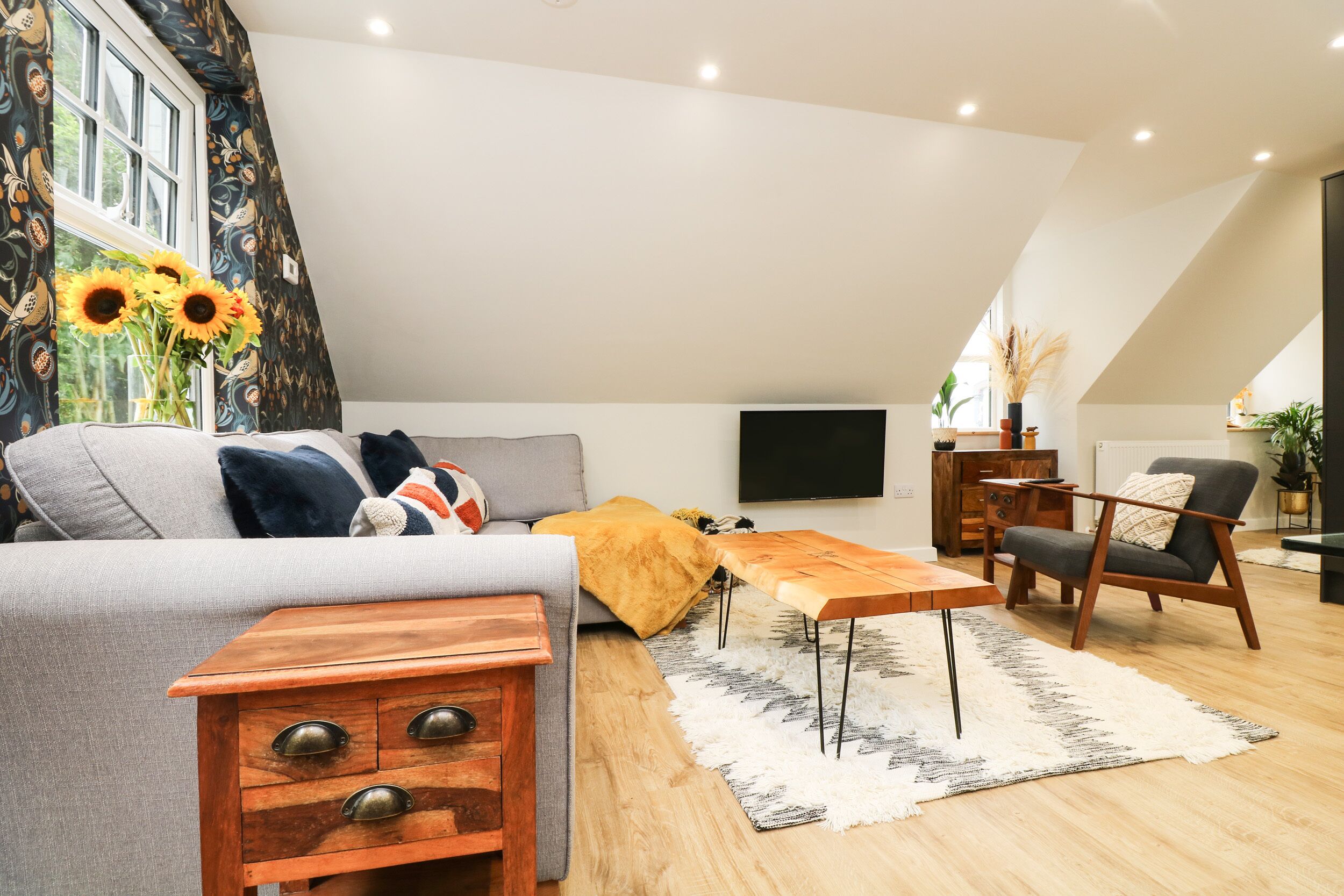 Een moderne woonkamer met een bank, een houten salontafel en een fauteuil. Er is een televisie aan de muur bevestigd en een raam met gordijnen.