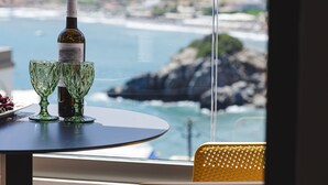 Ein Tisch mit einer Flasche und zwei Gläsern, ein gelber Stuhl und Blick auf das Meer und Felsen durch ein Fenster.