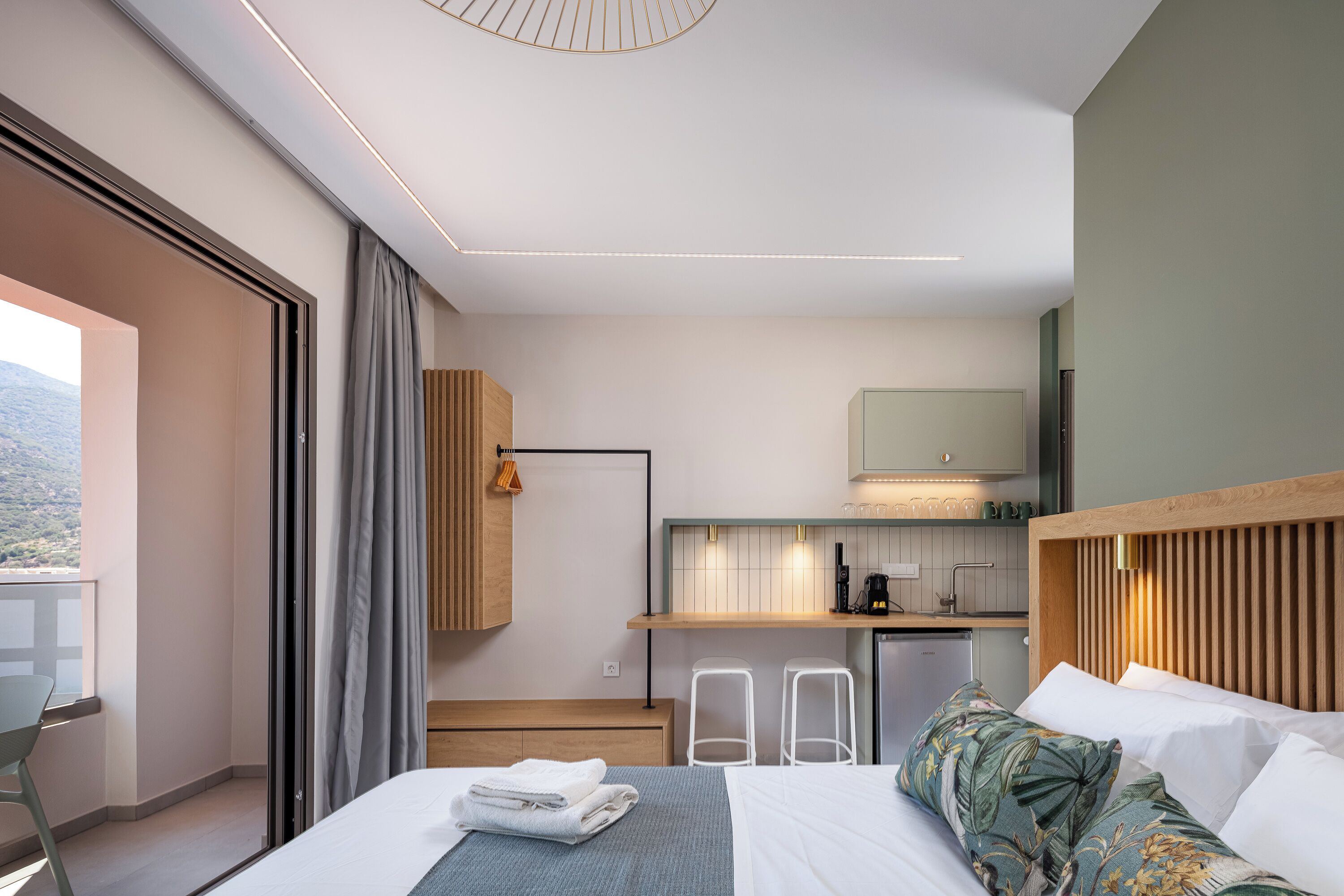 A modern hotel room with a bed, a small kitchenette, a balcony, and a view of the outdoors.