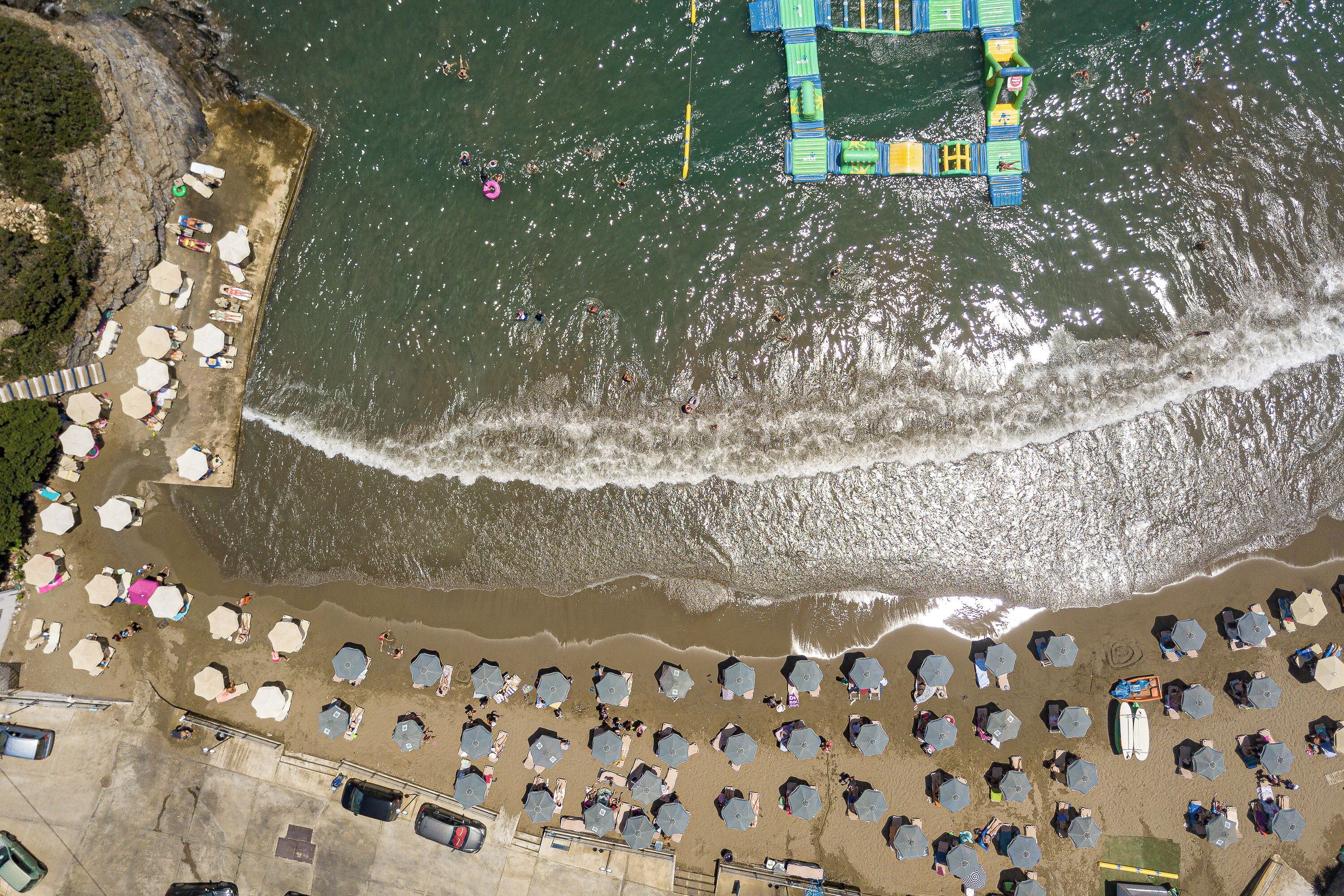 An aerial view of a beach with numerous umbrellas and sun loungers arranged in rows, a lifeguard tower, and a small pier extending into the water.