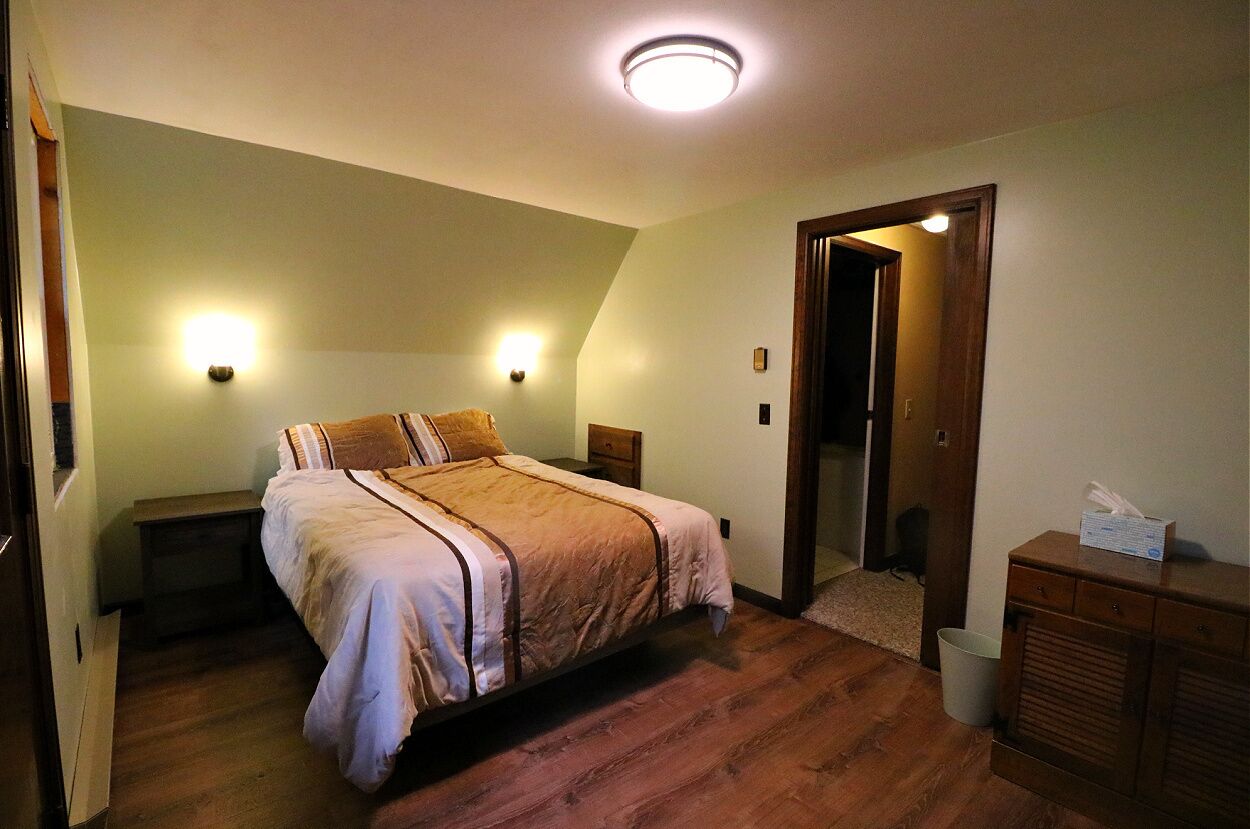 Ein Schlafzimmer mit einem Bett, Nachttischen, einem Beistelltisch, einer hölzernen Kommode und einer Tür zu einem anderen Raum.