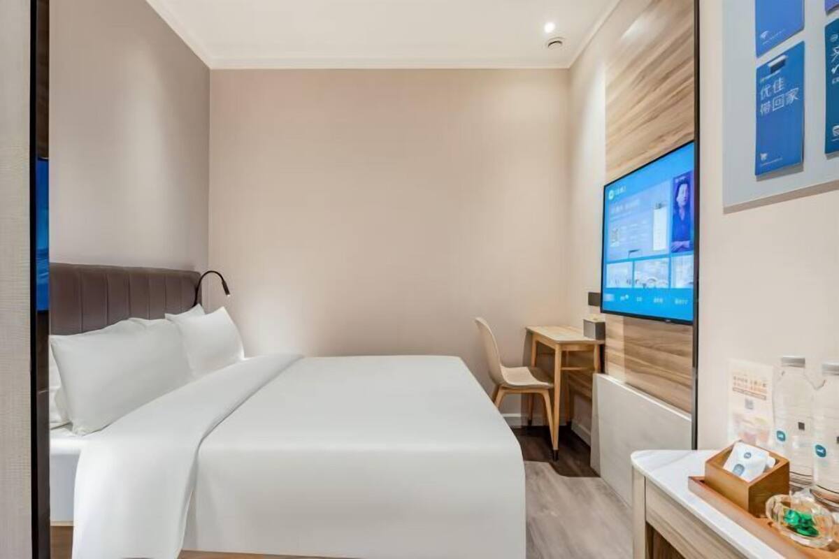 A hotel room with a bed, a desk, a chair, a TV mounted on the wall, and a small table with items on it.