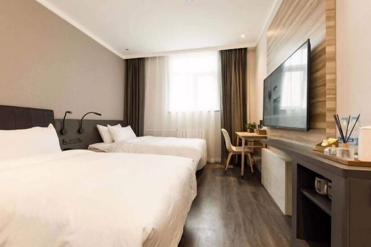 A hotel room with two beds, a desk, a chair, a flat-screen TV, and a large window with curtains.