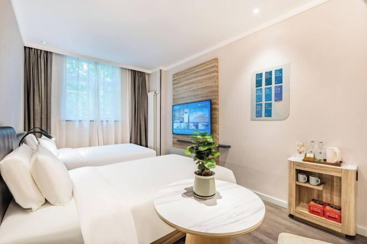 A hotel room with a bed, a round table with a plant, a TV on the wall, and a small cabinet with items.