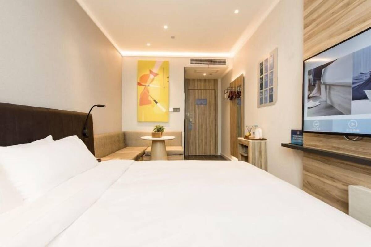 A hotel room with a bed, a small table with a plant, a TV on the wall, and a wooden panel design.