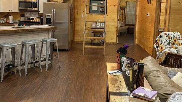 Un interior de cabaña acogedor con detalles de madera, una cocina con electrodomésticos modernos, un comedor con taburetes y una sala de estar con un sofá y una escalera que conduce a un altillo.