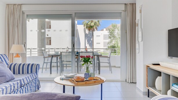 A modern living room with a sofa, a coffee table with pastries, a TV, and a balcony with a table and chairs. - First floor apartment in Bellresguard complex, with pool, near the beach (Puerto Pollensa)