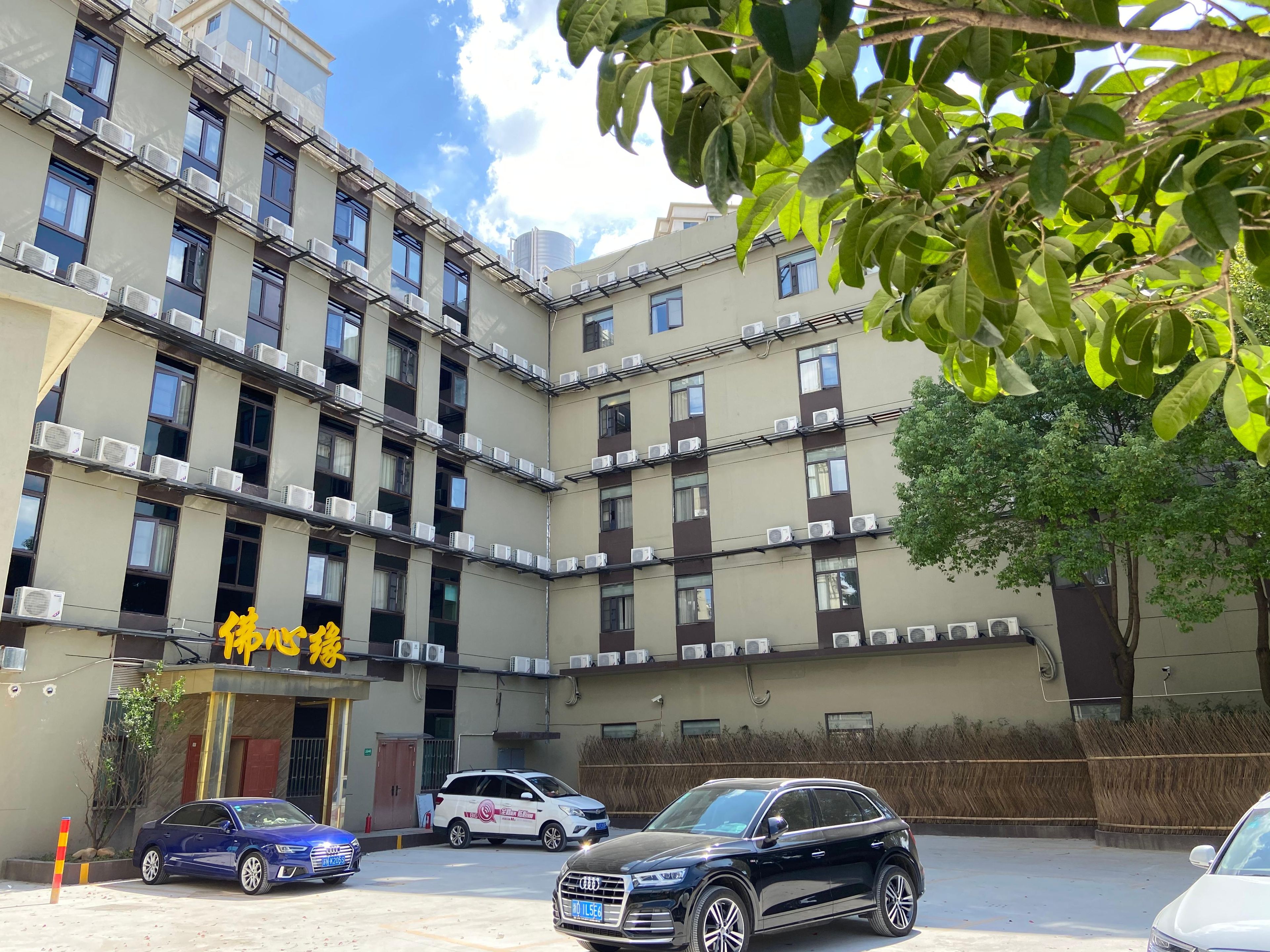 A multi-story building with air conditioning units, a parking area with several cars, and a tree in the foreground.