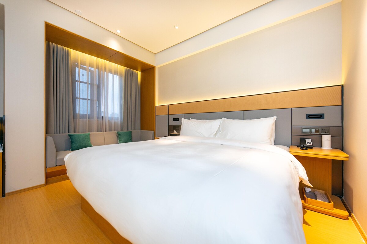 A modern hotel room with a large bed, a bedside table with a phone and a lamp, and a window with curtains.