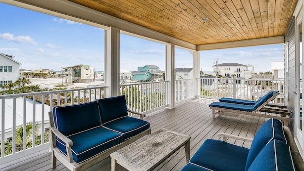 A covered outdoor deck with wooden flooring, a wooden ceiling, and white railings. There are blue cushioned seating areas and a small table. The deck overlooks a beach and houses. - Luxury Gulf View Home with Private Pool! Steps to the beach! (Pensacola Beach)