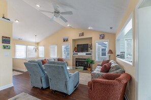 A living room with a fireplace, a ceiling fan, and a large window. - High Cotton - Old Nags Head Place, Ocean View, 325 Yds to Beach, Elevator, Pool, Hot Tub, Tiki Bar (Nags Head)