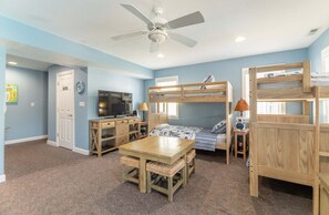 A room with bunk beds, a ceiling fan, a television, and a table with chairs. - High Cotton - Old Nags Head Place, Ocean View, 325 Yds to Beach, Elevator, Pool, Hot Tub, Tiki Bar (Nags Head)