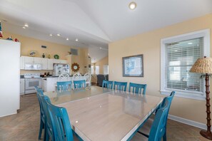 A dining area with a glass table and blue chairs, a kitchen with white appliances, and a window with blinds.