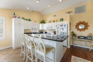A kitchen with white cabinetry, a central island with chairs, a microwave, and a refrigerator with magnets. - High Cotton - Old Nags Head Place, Ocean View, 325 Yds to Beach, Elevator, Pool, Hot Tub, Tiki Bar (Nags Head)