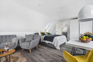 Une chambre moderne avec un lit, deux fauteuils gris, une table basse en bois, un bureau blanc avec un panier de fruits et une grande fenêtre.