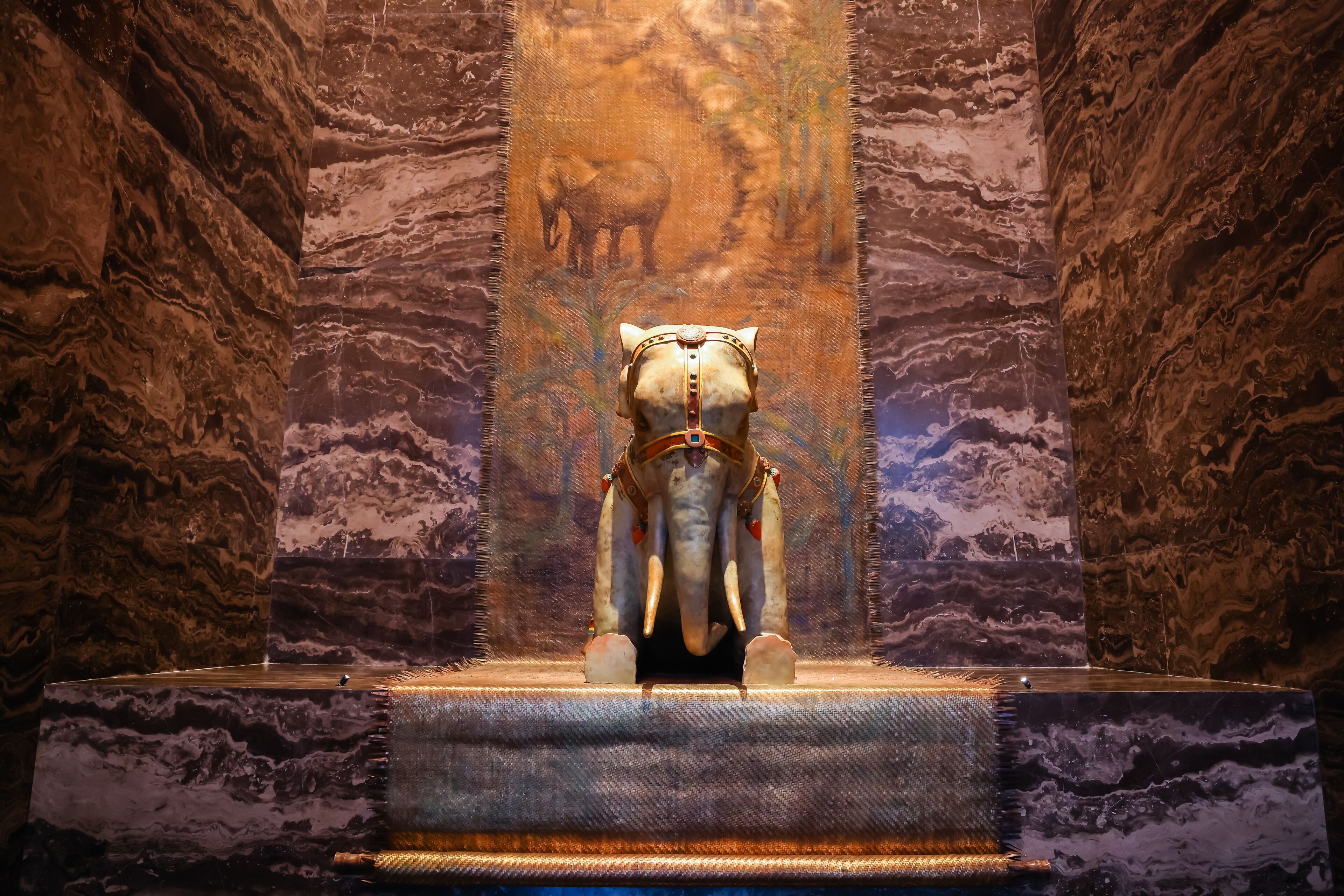 Una estatua de un elefante en un museo, con paredes de mármol y un cuadro vertical al fondo.