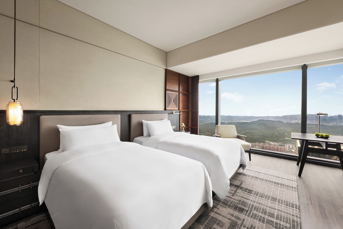 A modern hotel room with two beds, a desk, and a large window offering a city view.