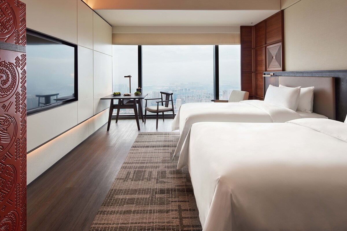 A modern hotel room with two beds, a dining table, and a city view.