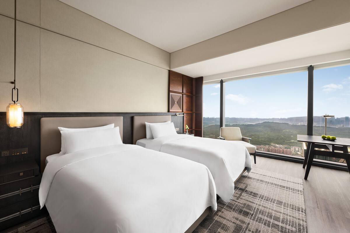 Una habitación de hotel moderna con dos camas, un escritorio y un amplio ventanal con vistas a la ciudad.