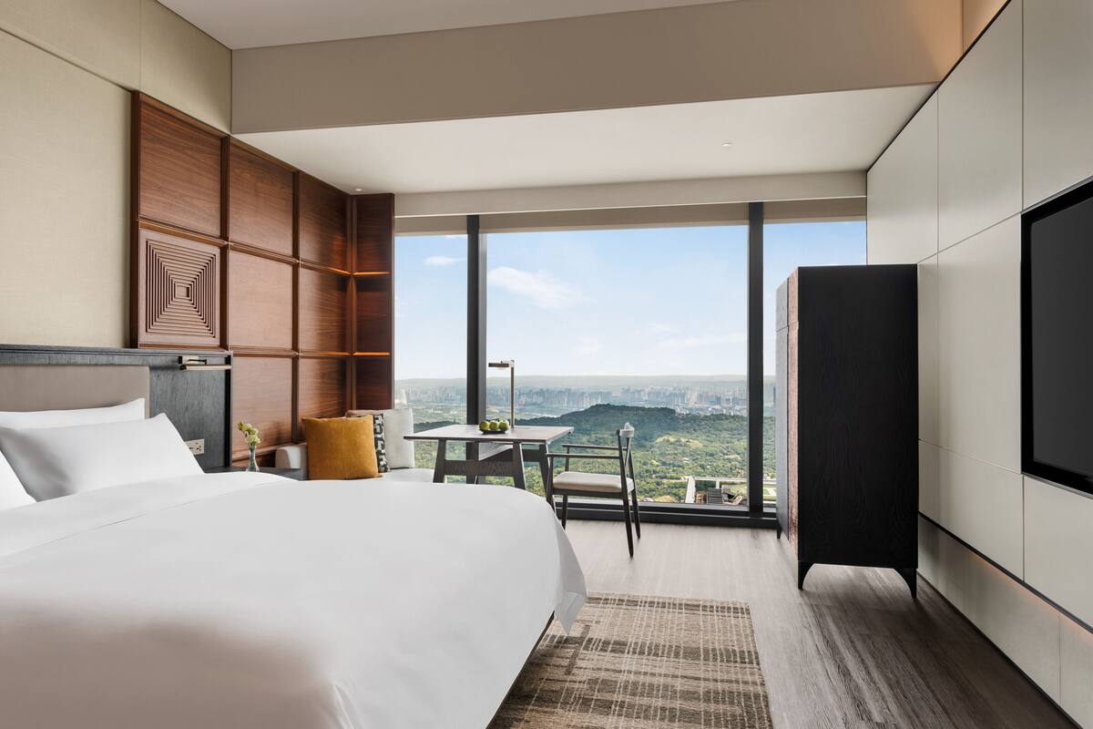 Una habitación de hotel moderna con una cama grande, un escritorio y vistas a un paisaje urbano a través de amplios ventanales.