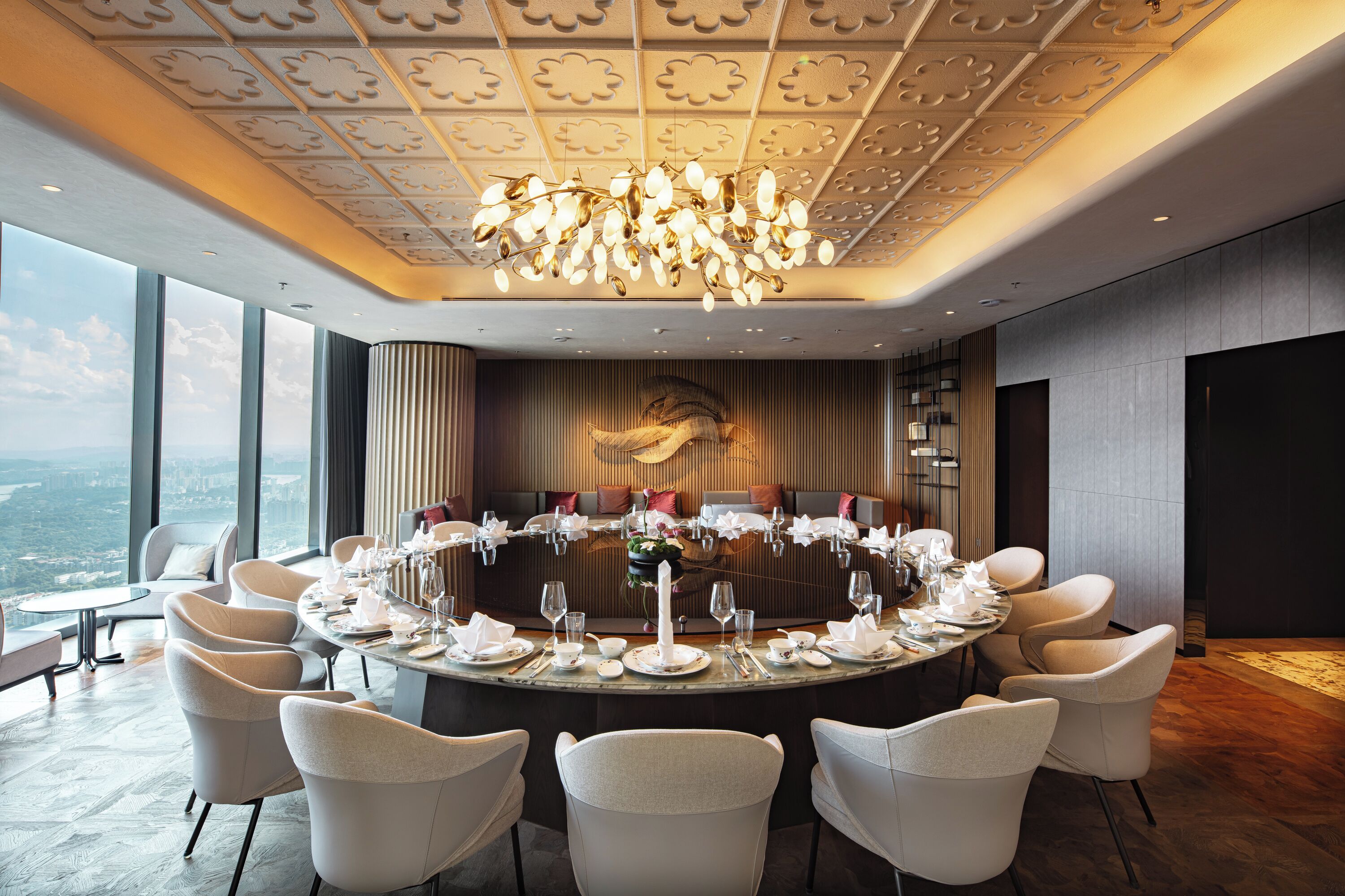 A round dining table set for a formal event, surrounded by white chairs, with a chandelier overhead and a city view through large windows.