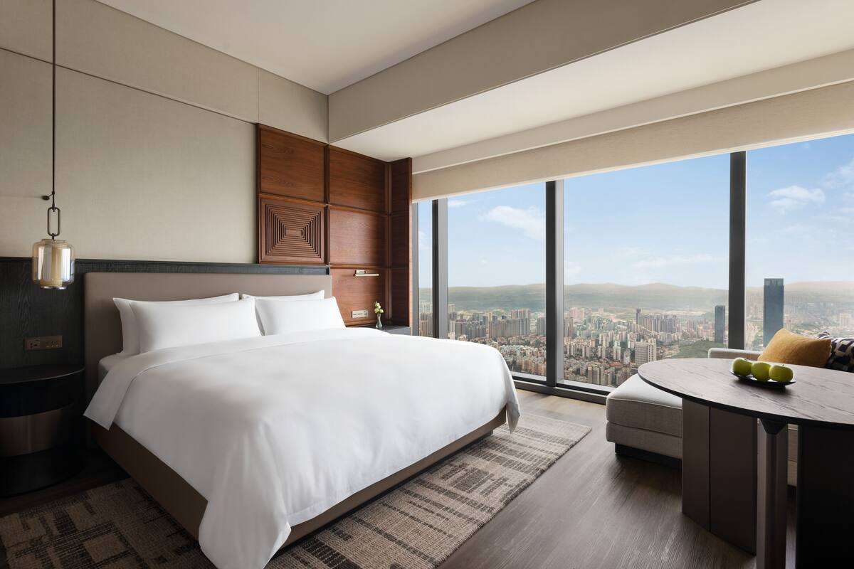 Habitación de hotel moderna con una cama grande, una mesa de comedor redonda y vistas a la ciudad a través de grandes ventanales.