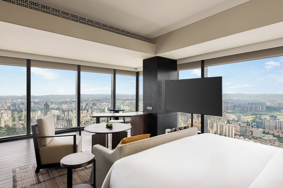 Una habitación de hotel moderna con una cama grande, un televisor montado en la pared, una zona de estar con una mesa y sillas, y vistas a la ciudad a través de amplias ventanas.